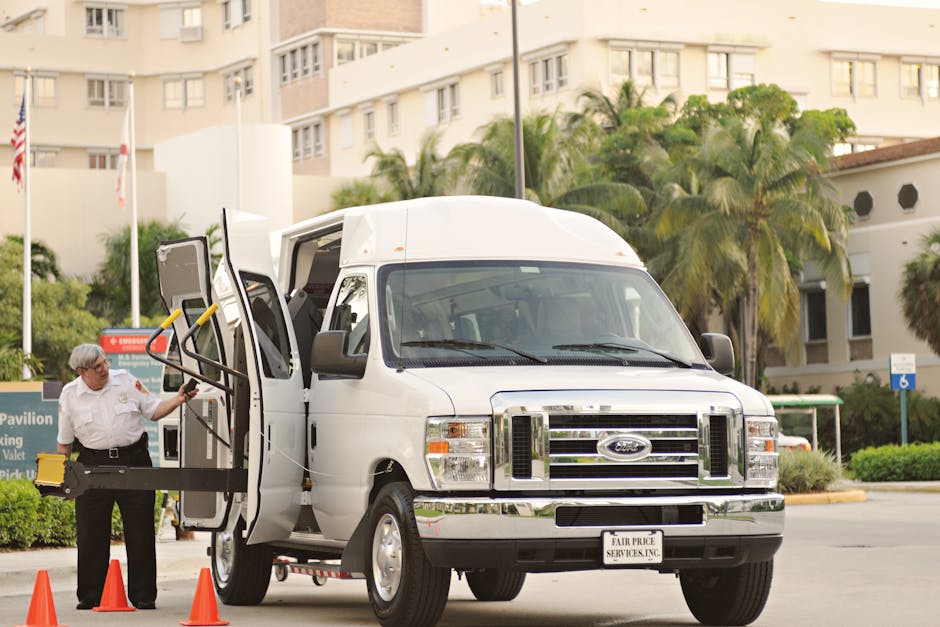 A white Ford transit van from Man with Van Gidea Park is parked outdoors on a paved area near a building with multiple floors, tall palm trees, and greenery in the background. The sliding side door of the van is open, revealing the interior designated for furniture transport and packing materials. A worker dressed in a white uniform, wearing glasses, is outside the van, holding a yellow and black hand truck or dolly, preparing to load or unload boxes wrapped in plastic film and packing materials, which are visible inside and near the entrance. The van's rear doors are closed, and there are orange safety cones on the pavement, indicating an active loading or home relocation process. The scene captures a professional moving operation or furniture transport, reflecting the logistical aspects involved in house removals or packing and moving services, supporting the content about best van access tips for Heath Drive terraces, Gidea Park.