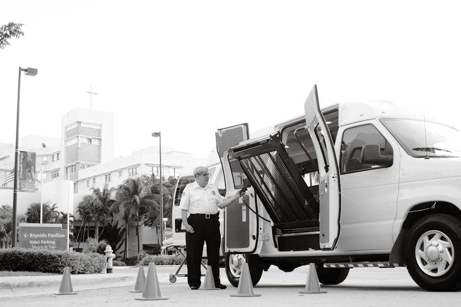 A man wearing glasses and a white shirt is standing next to a white van with its side door open, revealing a large loading ramp and interior space prepared for furniture transport during a home relocation. He appears to be securing or inspecting the loading process, which is taking place on a paved area marked by orange traffic cones. In the background, there are tall buildings, palm trees, and streetlights, indicating an urban setting near Gidea Park. The scene is well-lit with natural daylight, capturing the detailed positioning of the van, packaging materials like blankets or protective covers, and the surroundings for facilitating furniture transport and packing in house removals, as managed by Man with Van Gidea Park.