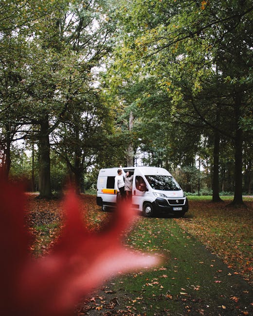 A white Ford transit van from Man with Van Gidea Park is parked outdoors on a paved area near a building with multiple floors, tall palm trees, and greenery in the background. The sliding side door of the van is open, revealing the interior designated for furniture transport and packing materials. A worker dressed in a white uniform, wearing glasses, is outside the van, holding a yellow and black hand truck or dolly, preparing to load or unload boxes wrapped in plastic film and packing materials, which are visible inside and near the entrance. The van's rear doors are closed, and there are orange safety cones on the pavement, indicating an active loading or home relocation process. The scene captures a professional moving operation or furniture transport, reflecting the logistical aspects involved in house removals or packing and moving services, supporting the content about best van access tips for Heath Drive terraces, Gidea Park.