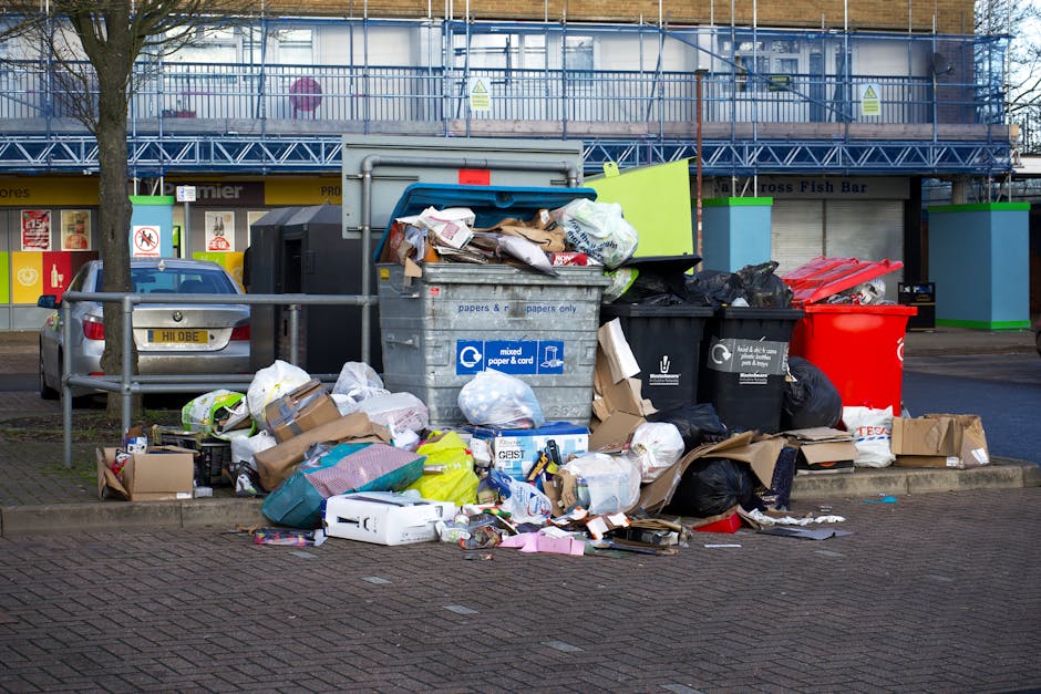A collection of overflowing rubbish bins and waste materials on a paved sidewalk outside a commercial building, including large grey mixed paper and card waste containers, black and red trash bins filled with various household and packaging rubbish, and scattered cardboard boxes, plastic bags, paper debris, and packaging materials on the ground. Behind the waste area, a metal railing separates the bins from parked vehicles, including a silver car, and a building with storefront signage and a blue scaffolding structure is visible in the background. The scene is lit with natural daylight, illustrating waste collection and disposal which may be relevant to household removal services provided by Man with Van Gidea Park during home relocation processes.