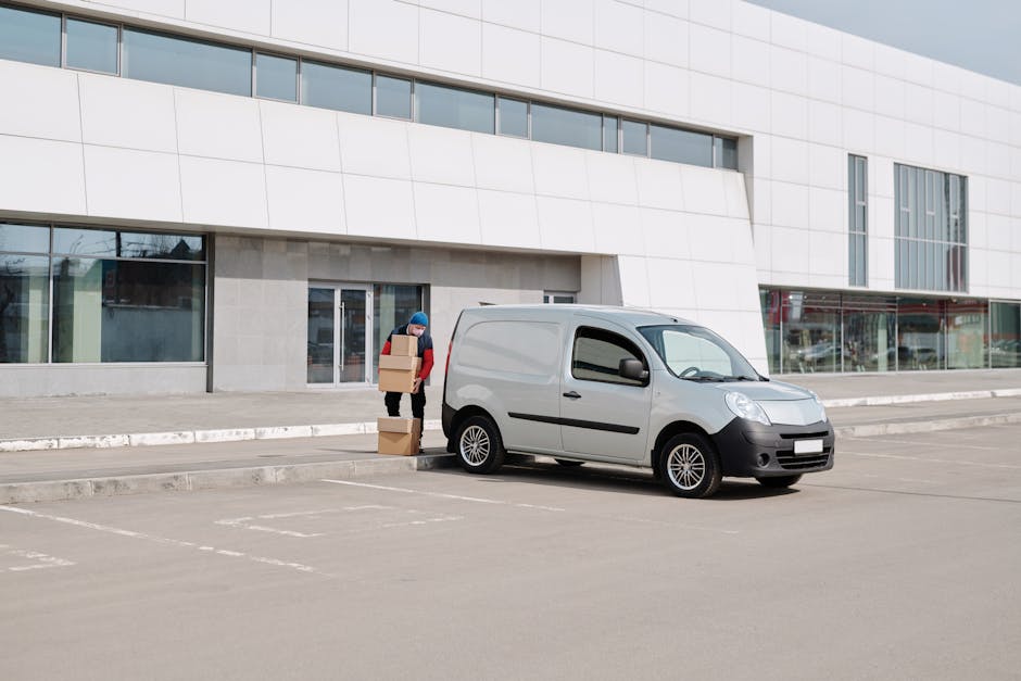 A person wearing a blue hat and red jacket standing beside a silver delivery van parked in an empty outdoor lot in front of a modern, multi-story commercial building. The person is holding three cardb