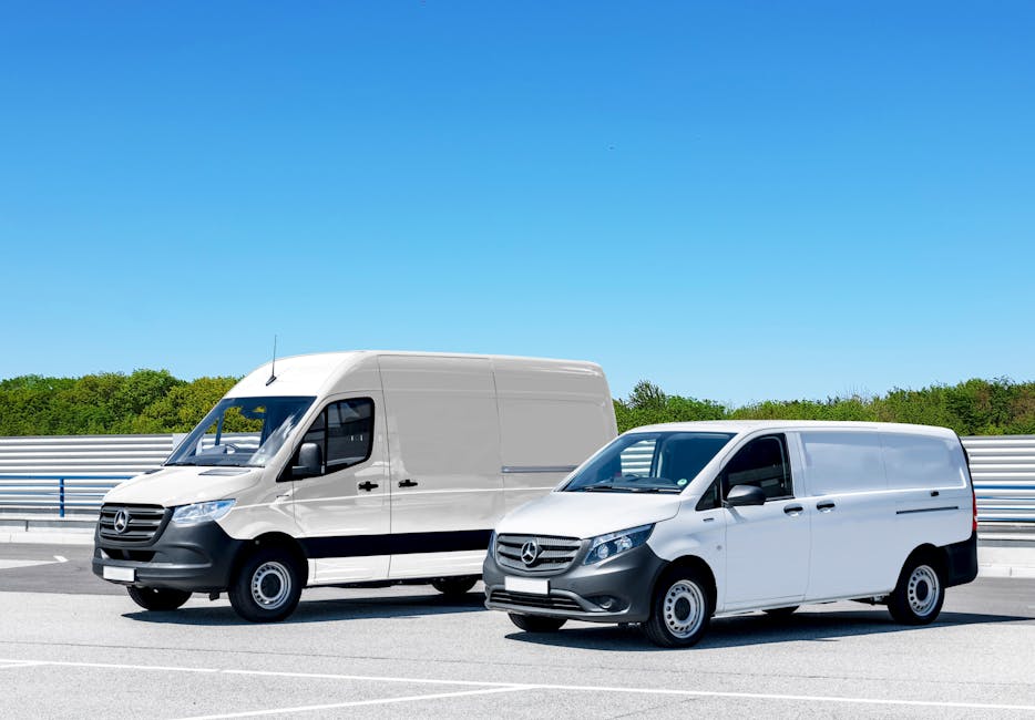 A person wearing a blue hat and red jacket standing beside a silver delivery van parked in an empty outdoor lot in front of a modern, multi-story commercial building. The person is holding three cardb