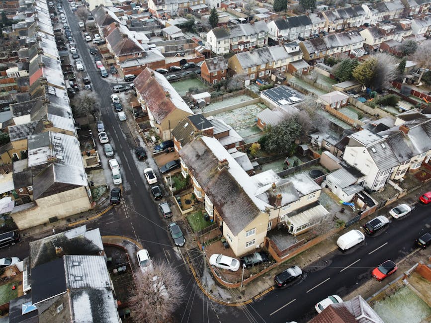 An aerial view of a residential neighborhood in Gidea Park during winter, showing multiple terraced houses and semi-detached homes with rooftops dusted in snow. Narrow streets are lined with parked cars, and some homes have small front gardens enclosed by low brick walls or fences. The image captures the compact layout characteristic of the area, with visible roadways and a few trees scattered throughout the neighborhood. This scene relates to home relocation and furniture transport, as it highlights the typical environment where house removals and moving services by Man with Van Gidea Park might take place, especially during seasonal packing and moving processes involving household items and vehicles likely used for home moves.