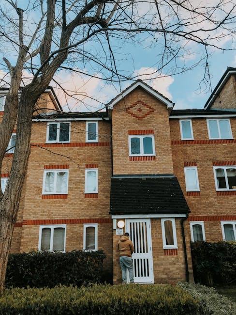 An individual wearing a brown jacket and grey trousers stands outside a three-storey brick residential building, which features a white front door with vertical slats and a black-tiled porch roof. The building has multiple white-framed windows and decorative red brickwork accents around the windows and a diamond-shaped pattern on the central gable. In front of the entrance, there are neatly trimmed bushes and a small lawn. Large, leafless trees with bare branches are visible overhead, and the sky is partly cloudy with patches of blue. This scene depicts a typical home relocation or moving process, likely organized by Man with Van Gidea Park as part of their removals service for residents in the Gidea Park area, focusing on moves and furniture transport logistics.