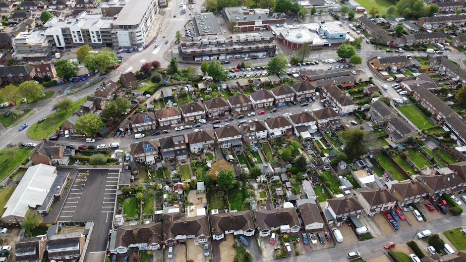 An aerial view of a residential neighborhood in Gidea Park during winter, showing multiple terraced houses and semi-detached homes with rooftops dusted in snow. Narrow streets are lined with parked cars, and some homes have small front gardens enclosed by low brick walls or fences. The image captures the compact layout characteristic of the area, with visible roadways and a few trees scattered throughout the neighborhood. This scene relates to home relocation and furniture transport, as it highlights the typical environment where house removals and moving services by Man with Van Gidea Park might take place, especially during seasonal packing and moving processes involving household items and vehicles likely used for home moves.