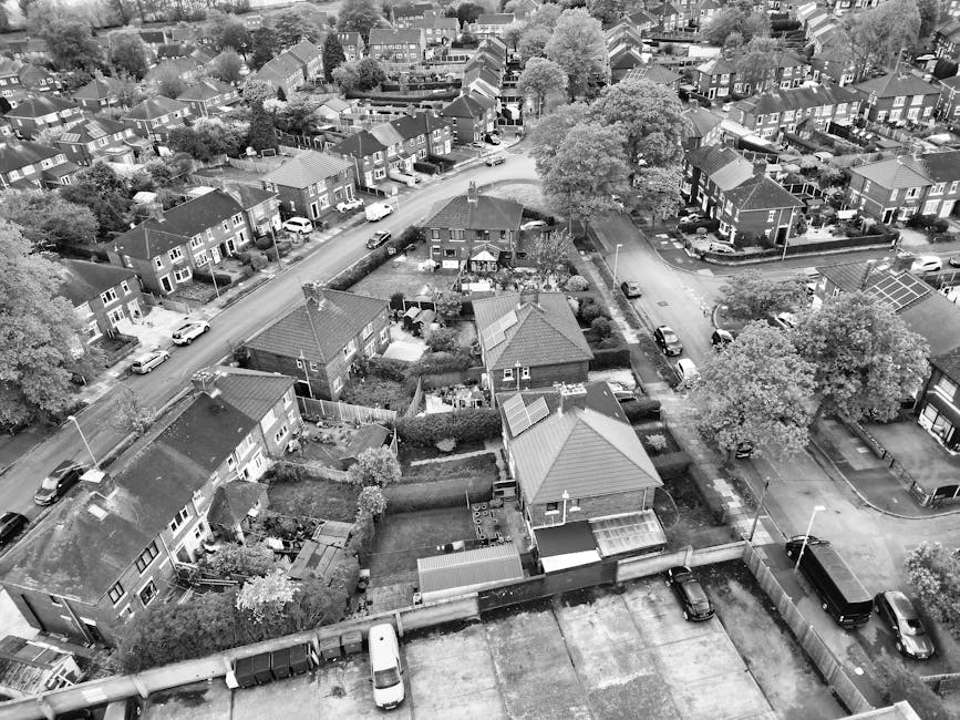 An aerial view of a residential neighbourhood with a mix of detached and semi-detached houses, trees, and parked cars along the streets and driveways. In the foreground, multiple houses with pitched roofs are visible, some with small gardens enclosed by hedges or fences. A driveway adjacent to one house contains a white van, likely used for home relocation purposes. Several cardboard boxes, wrapped furniture, and packing materials are seen outside the houses, indicating a packing and moving process. Near a gated entrance, a man with a van is engaged in loading or unloading furniture wrapped in plastic and blankets onto the vehicle. The scene is well-lit with natural daylight, capturing the spatial arrangement of properties, vehicles, and outdoor items, as typical during a house move coordinated by a professional removals company like Man with Van Gidea Park, supporting efficient furniture transport and house relocation services in the Gidea Park area.