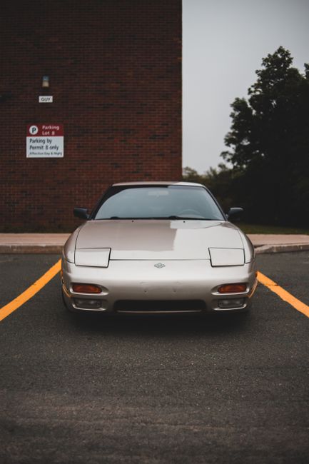 A silver sports car with a sleek, low-profile design is parked in a marked parking space in front of a red brick building during daytime. The car features a smooth, aerodynamic shape with pop-up headlights in the closed position, a small Nissan emblem on the front hood, and black side mirrors. The parking space is outlined with yellow lines, and the surface is paved with asphalt. Behind the car, the brick building has a small sign indicating parking restrictions, and to the right, there are trees and greenery visible under an overcast sky. The scene reflects a quiet, urban environment suitable for home relocation or furniture transport logistics, with the vehicle positioned for easy access to a nearby property. Man with Van Gidea Park may utilize such vehicles in their removals and moving services, including packing and loading processes within this setting.