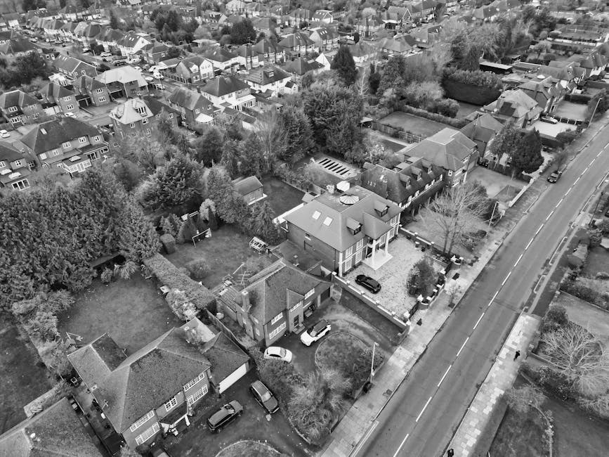 An aerial view of a residential neighbourhood with a mix of detached and semi-detached houses, trees, and parked cars along the streets and driveways. In the foreground, multiple houses with pitched roofs are visible, some with small gardens enclosed by hedges or fences. A driveway adjacent to one house contains a white van, likely used for home relocation purposes. Several cardboard boxes, wrapped furniture, and packing materials are seen outside the houses, indicating a packing and moving process. Near a gated entrance, a man with a van is engaged in loading or unloading furniture wrapped in plastic and blankets onto the vehicle. The scene is well-lit with natural daylight, capturing the spatial arrangement of properties, vehicles, and outdoor items, as typical during a house move coordinated by a professional removals company like Man with Van Gidea Park, supporting efficient furniture transport and house relocation services in the Gidea Park area.