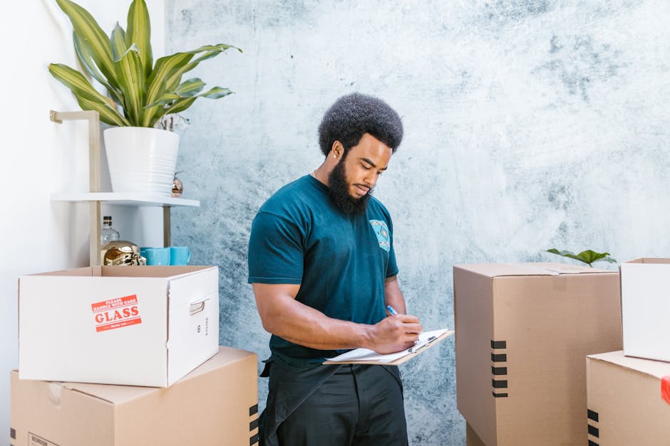 A man wearing a yellow t-shirt and blue jeans is standing indoors amidst several open cardboard boxes and some closed ones stacked in the background, indicating a home relocation or moving process. He is holding a clipboard in his left hand and using a black marker in his right hand to mark or record details, likely related to packing or inventory. The boxes are empty or filled with packing materials, and the scene is set in a bright room with light-colored walls, a hardwood floor, and a potted plant with broad green leaves placed beside the boxes. This image captures the typical activities of packing, preparing for furniture transport, or organizing items during a house move, as done by professional removals services like Man with Van Gidea Park, supporting thorough and efficient moving logistics.