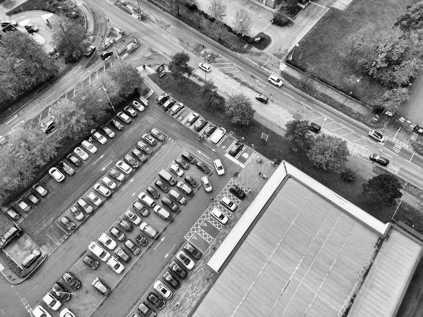 A black-and-white aerial photograph showing a large, multi-storey parking lot filled with numerous parked cars arranged in neat rows, situated adjacent to a building with a flat roof. Surrounding the parking area are trees, sidewalks, and roads with moving vehicles. In the foreground, the roof of a building is visible, with a portion of its edge aligned with the parking lot below. The photograph captures a scene relevant to house removals and relocation services, illustrating logistical considerations such as parking and loading zones, essential for planning efficient furniture transport and home relocation processes. The image is associated with Man with Van Gidea Park, a company providing removals, and emphasizes the importance of understanding parking and loading challenges during Gidea Park moves, as highlighted in the page titled 'Parking and loading challenges for Gidea Park moves.'