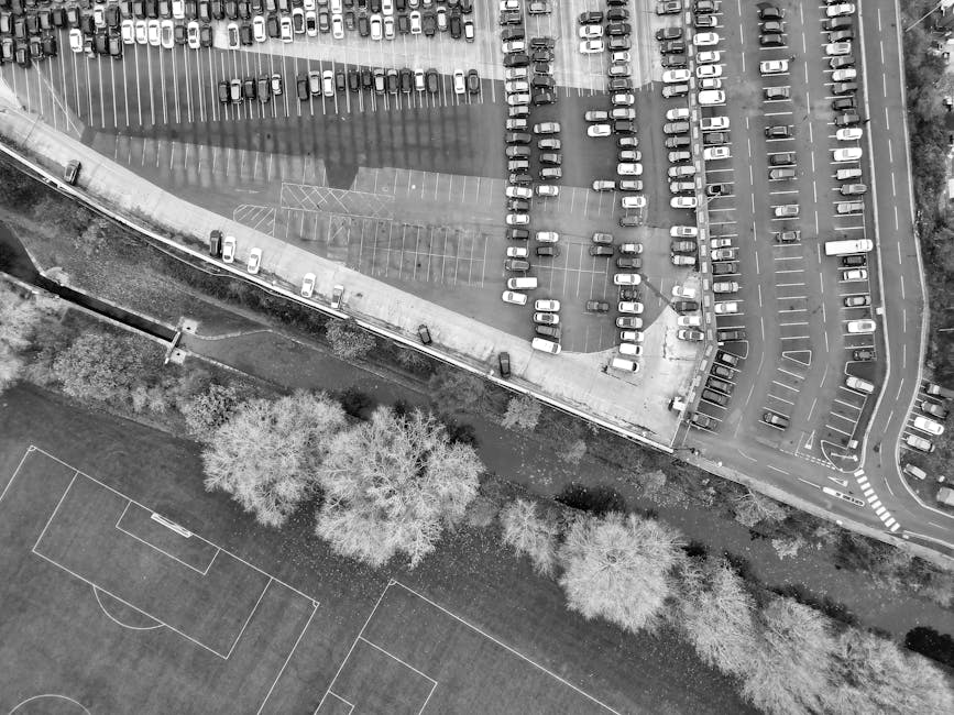 An aerial black-and-white photograph showing a large parking area adjacent to a building, which is being used for loading and unloading furniture and household items during a home relocation. Multiple vehicles are parked in designated spaces, with some positioned near the building's entrance, facilitating the loading process. Several large cardboard boxes and furniture pieces covered in protective fabric or plastic wrap are visible inside or near the building's doorway, with some being carried by movers. A trolley and moving straps are also present near the entrance, indicating active loading activity. The parking area is partially occupied, with clear markings and pathways for vehicle movement. Beyond the parking lot, there is a sports field with visible boundary lines and trees lining the perimeter, emphasizing the outdoor environment involved in the relocation process. The scene highlights the logistical challenges of parking and loading for house removals, as managed by Man with Van Gidea Park, within a typical urban setting.