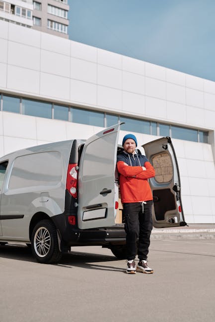 A man wearing a blue beanie and a red jacket stands outside a grey cargo van parked adjacent to a modern white building with large windows. The van's rear doors are open, revealing an empty cargo spac