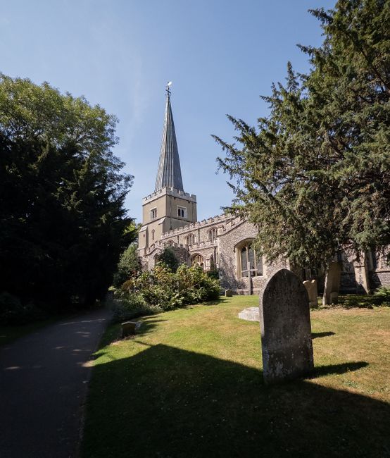 A narrow paved pathway leading towards a historic church with a tall, pointed steeple under a bright blue sky, surrounded by lush green trees and grass. Several weathered gravestones are visible in the churchyard, with one prominent upright headstone in the foreground casting a shadow on the grass. The church building features stone walls with arched windows, and the scene suggests an outdoor setting during daytime. The environment is calm and well-maintained, indicative of a peaceful churchyard area often associated with house removals or relocation services, as seen on the GIDEA PARK website.