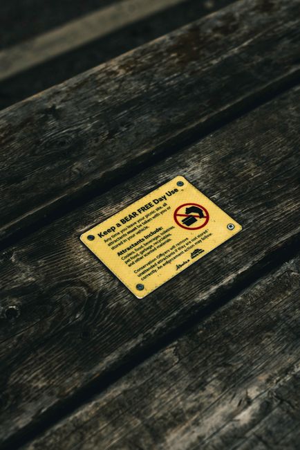 Close-up image of a yellow warning label attached to a weathered wooden surface, likely a bench or table, with black screws securing it. The label instructs to keep the area bear-free and for daylight use only, featuring a symbol of a bear with a line through it. The background shows an outdoor environment with natural lighting, where the wooden surface appears aged with visible grain and texture. This image may relate to outdoor furniture used during home relocation or packing activities, emphasizing safety and regulatory compliance typical of house removals companies like Man with Van Gidea Park involved in furniture transport and moving logistics.