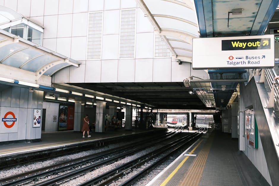 The image shows the exterior of a London Underground station at Hammersmith, with the station logo visible on a panel on the wall. The platform is partially covered by a curved, transparent roof structure, and has a yellow tactile paving strip near the edge for safety. There are a few passengers, including a person walking towards the stairs and a woman standing further down the platform. Multiple train tracks run through the station, curving slightly as they extend into the distance. To the right, an entrance/exit sign indicates the way out towards buses on Talgarth Road, with black and yellow signage. The surroundings include modern station architecture with metal cladding and windows, and the lighting is a combination of natural light entering from the entrance and artificial illumination along the platform. This scene depicts a typical urban rail station environment in London, suitable for documenting aspects of home relocation and moving process logistics involving public transport links.