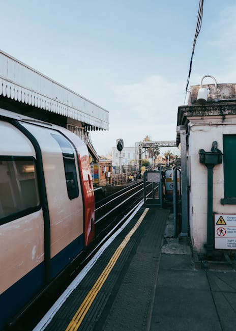 A view of a Gidea Park train station platform with a modern commuter train stopped alongside. The train has a sleek, curved exterior with a predominantly white and orange color scheme, featuring large, dark tinted windows. The platform border includes tactile paving with yellow lines for safety, and there are various objects such as stacked cardboard boxes, blankets, and packing materials for a home relocation visible closer to the station building. The station's building on the right features a white brick facade with green window shutters, a black mailbox, and safety signage. Overhead, electrical wires stretch across the sky, and the station setting is well-lit with natural daylight, indicating a clear day. Inside the station, there are trolleys and equipment used for furniture transport and packing during a house move coordinated by Man with Van Gidea Park, supporting efficient removal and moving services based on the scene depicted.