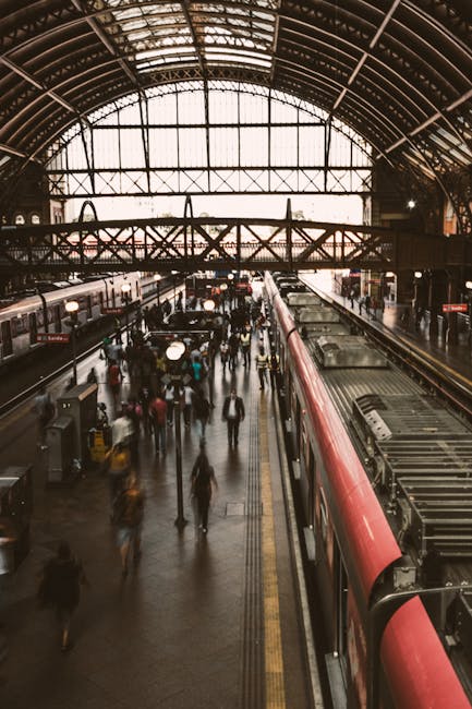 The image shows the exterior of a London Underground station at Hammersmith, with the station logo visible on a panel on the wall. The platform is partially covered by a curved, transparent roof structure, and has a yellow tactile paving strip near the edge for safety. There are a few passengers, including a person walking towards the stairs and a woman standing further down the platform. Multiple train tracks run through the station, curving slightly as they extend into the distance. To the right, an entrance/exit sign indicates the way out towards buses on Talgarth Road, with black and yellow signage. The surroundings include modern station architecture with metal cladding and windows, and the lighting is a combination of natural light entering from the entrance and artificial illumination along the platform. This scene depicts a typical urban rail station environment in London, suitable for documenting aspects of home relocation and moving process logistics involving public transport links.